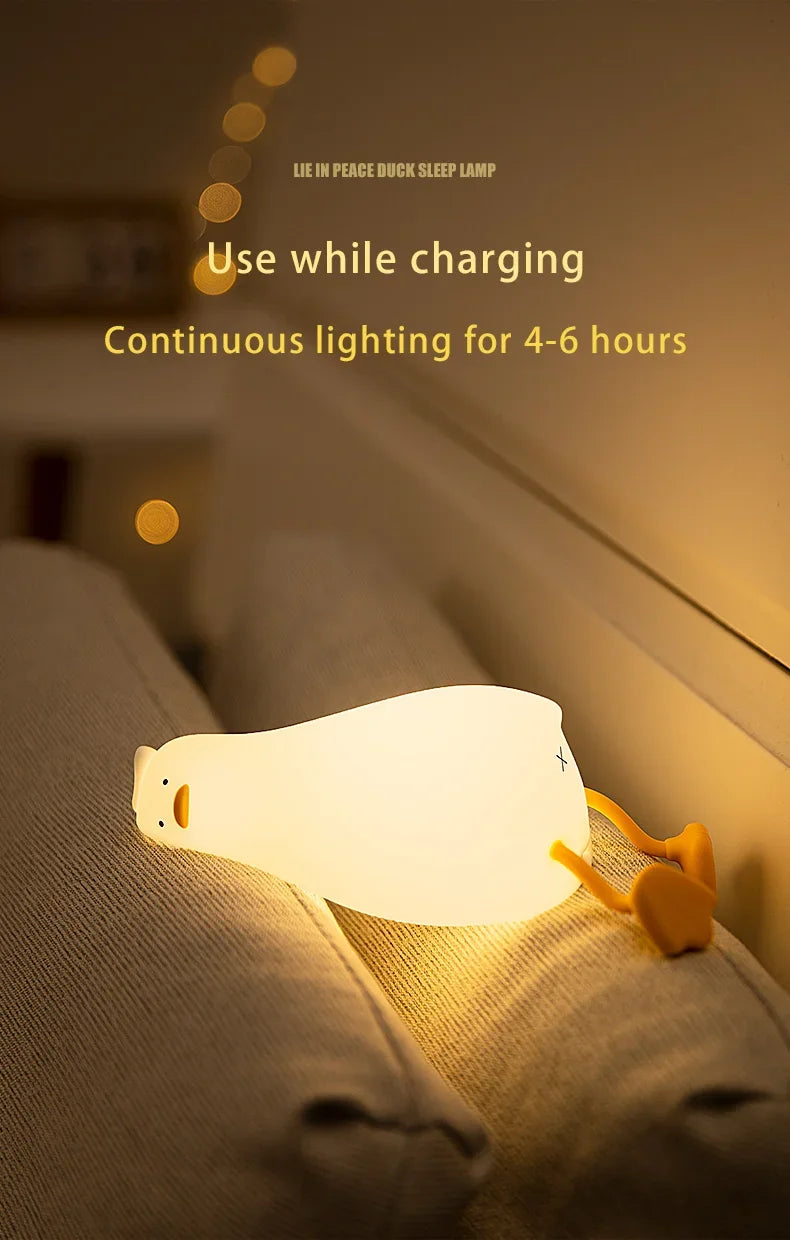 A white silicone duck touch night light is illuminated on a bed, used while charging.