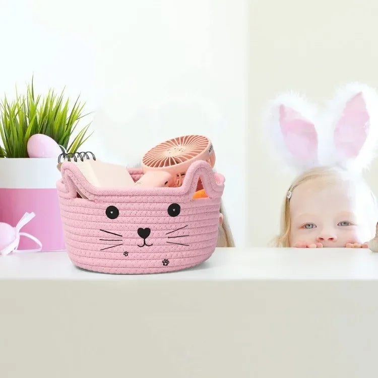 A pink woven cotton rope storage basket shaped like a cat is shown next to a baby with bunny ears.