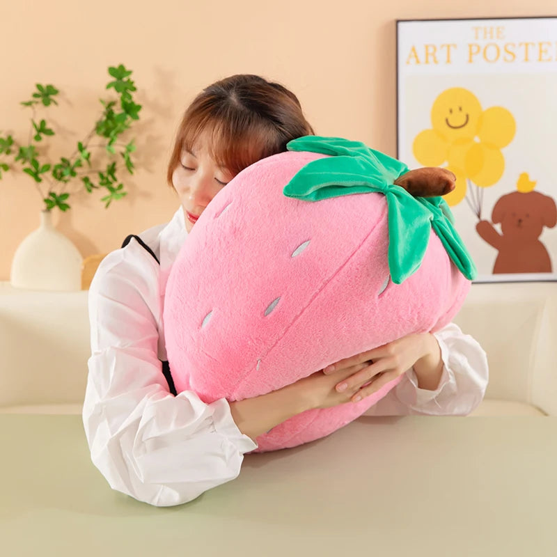 A person is holding a pink strawberry plush throw pillow with green leaves and a brown stem.