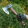 A hand-forged Viking tomahawk axe with a wooden handle and dark green leather grip is held in a natural outdoor setting.
