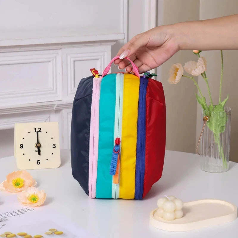 Eine farbenfrohe, leichte Reise-Toiletttasche mit Streifen in Blau, Grün, Gelb und Rot wird von einer Hand auf einer weißen O