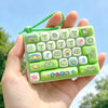 A hand holds a green cartoon keycap fidget keychain with various character and number keys.