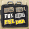 A black tactical patch with embroidered labels for MEDIC, FBI, RRT, and DEA on a beige fabric background.