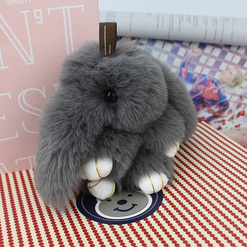 A fluffy gray bunny keychain with a pom pom tail resting on a red and white patterned surface.