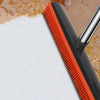 A black rubber broom and carpet rake with an orange bristles and silver adjustable handle resting on a white surface.