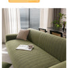 A textured olive green quilted stretch sofa cover displayed indoors with natural light and vertical blinds.