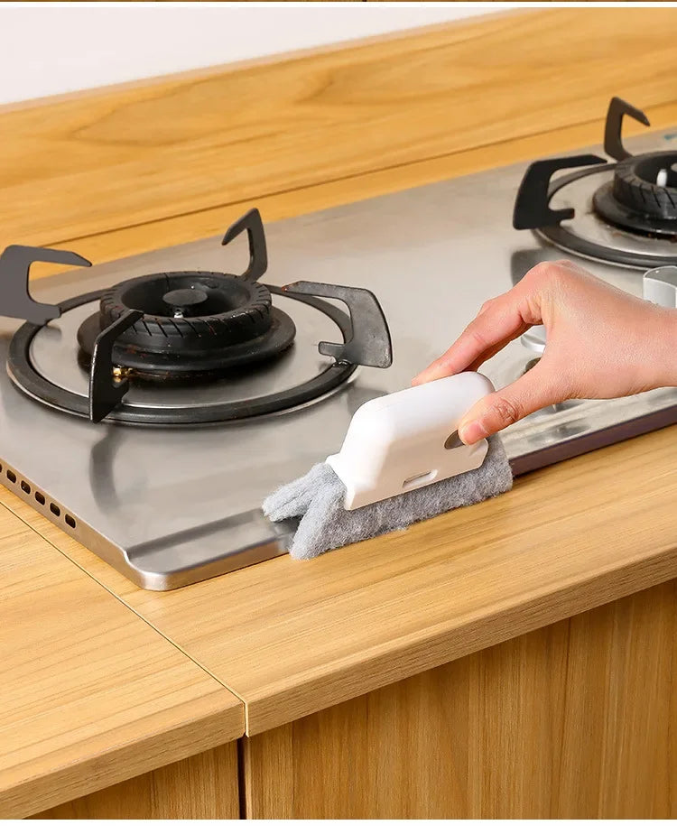 A hand is cleaning a stovetop burner using a white groove cleaning brush with a replaceable scouring head.
