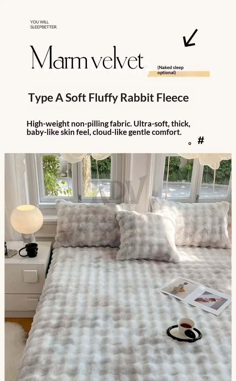 A light gray rabbit fleece blanket and pillows displayed on a bed, with natural light coming through the window.