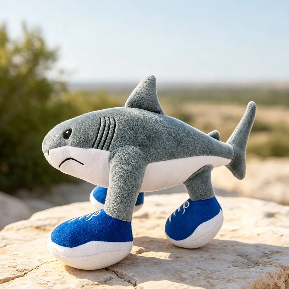 A gray and white plush toy of a shark with blue feet sitting on a light-colored rock.