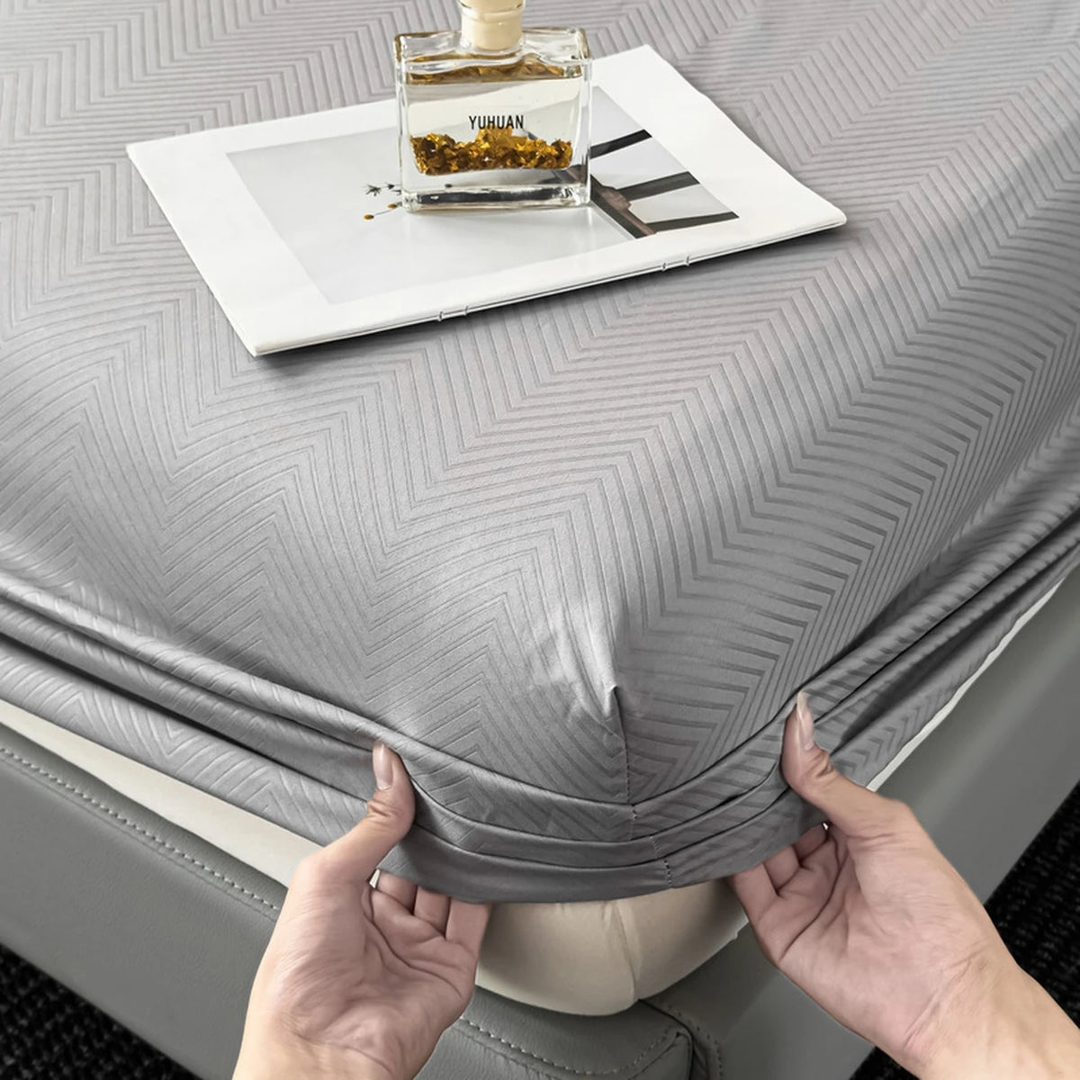 A gray brushed microfiber fitted sheet is shown on a bed, with a glass bottle of perfume resting on top.