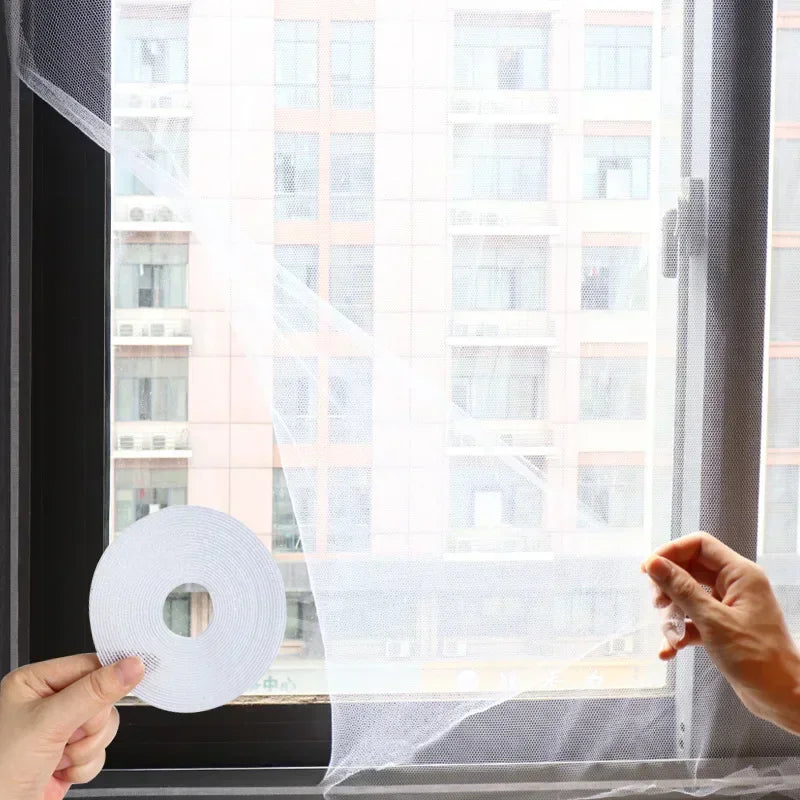 A hand holds a roll of white self-adhesive mosquito net material in front of a window.