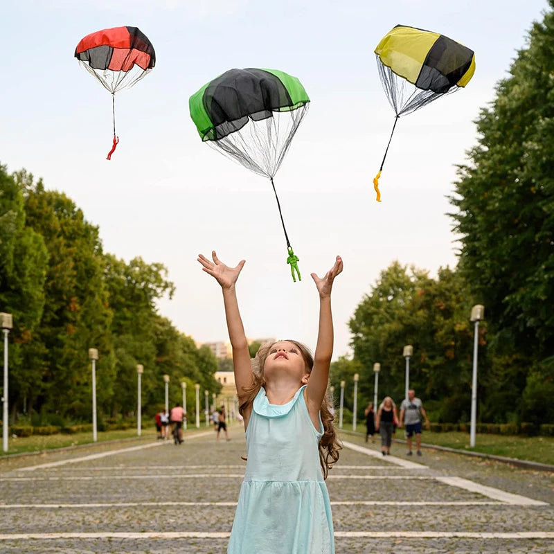 A young girl in a light blue dress is joyfully raising her arms while colorful hand-thrown parachute toys float above her in 