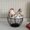A white ceramic hen-shaped egg basket with a lid, featuring painted details and a black wire base.