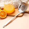 A stainless steel fine mesh skimmer spoon is shown next to a glass of orange juice and a slice of orange on a wooden board.