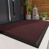 A dark red patterned non-slip doormat placed on a light gray floor near a wooden wall and potted plants.
