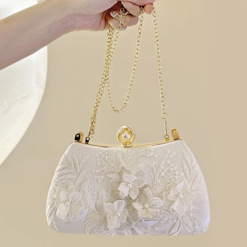 A white pearl embroidered evening clutch with a gold chain strap being held by a hand.