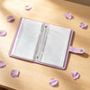 A lilac organizer binder with transparent pages is displayed on a wooden surface surrounded by pink flower petals.