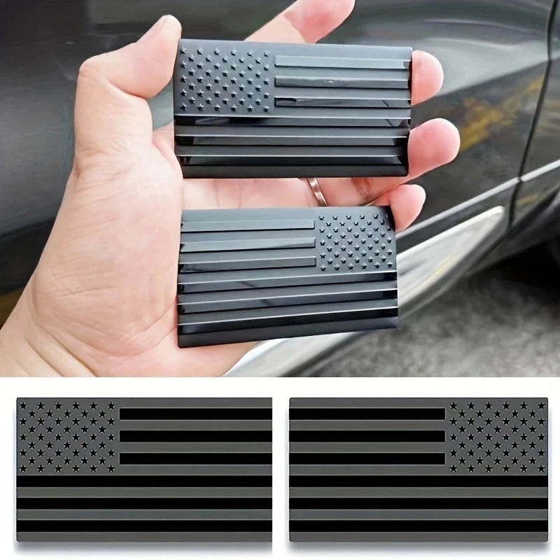 A hand holds a set of matte black 3D American flag car emblems featuring stars and stripes.