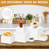 Six different sizes of white acrylic display risers stacked on a wooden table with various items.
