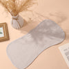 A beige baby burp cloth is displayed on a light pink background next to dried floral arrangements and a picture frame.
