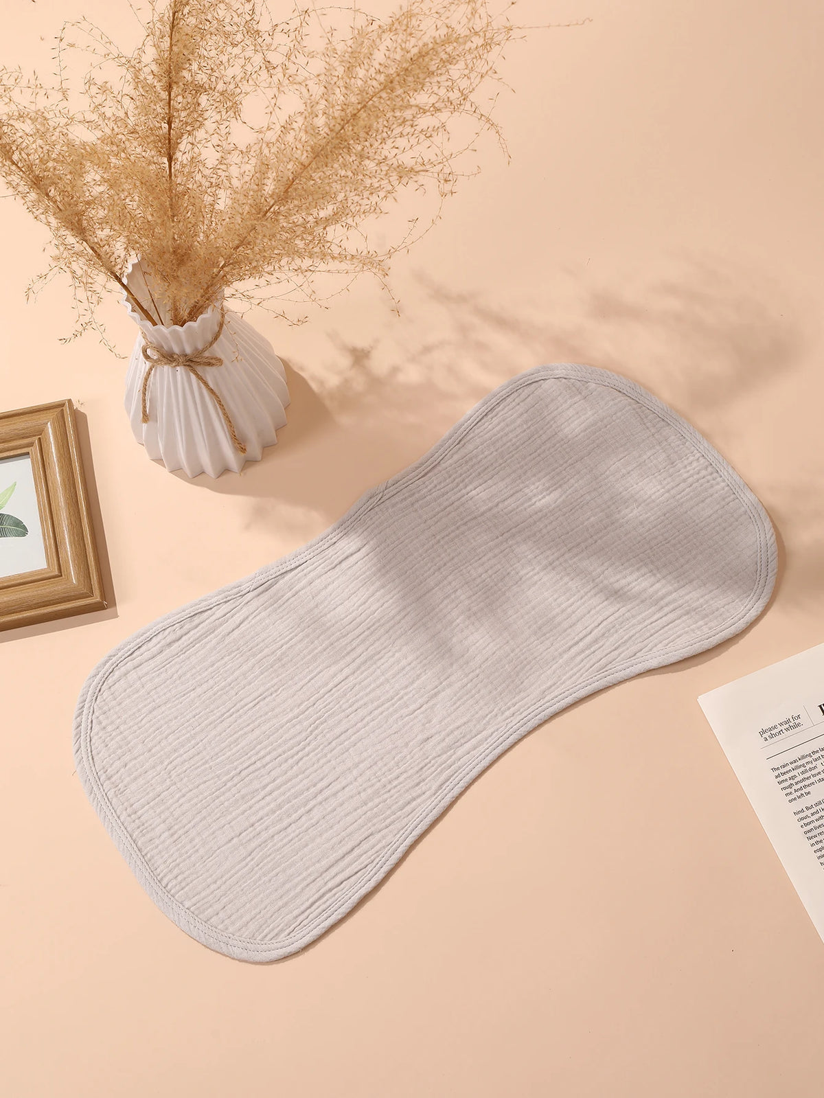 A beige baby burp cloth is displayed on a light pink background next to dried floral arrangements and a picture frame.