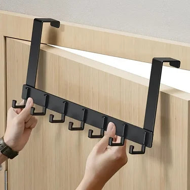 Hands installing a black over-the-door coat rack with six hooks onto a wooden cabinet door.