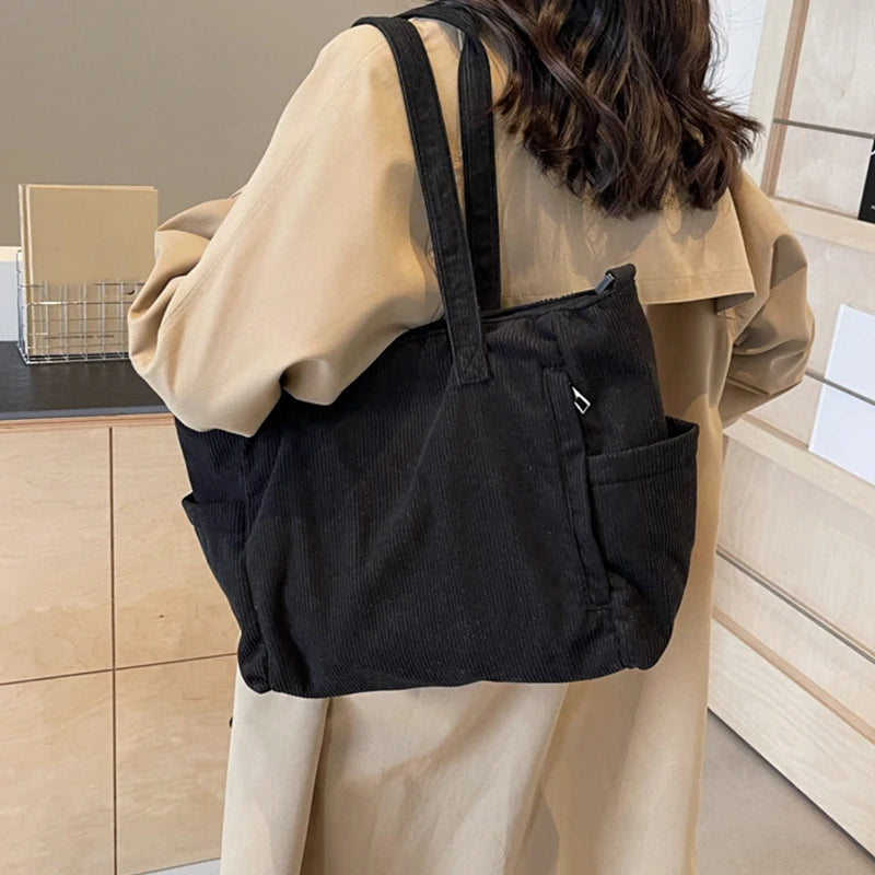 A woman is holding a black corduroy tote bag with an adjustable strap against a light beige coat.