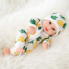 A baby doll wearing a white jumpsuit outfit with a yellow and green fruit pattern, lying on a white fluffy surface.