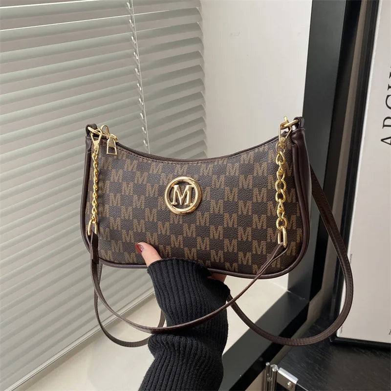 A brown PU leather crossbody bag with a gold metal M accent is held by a hand against a light background.