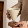 A hand holds a light beige ceramic coffee mug with a conical shape above a wooden base and spoon.