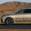 A silver car with a leopard print decal strip driving on a road under a dry, hilly landscape.