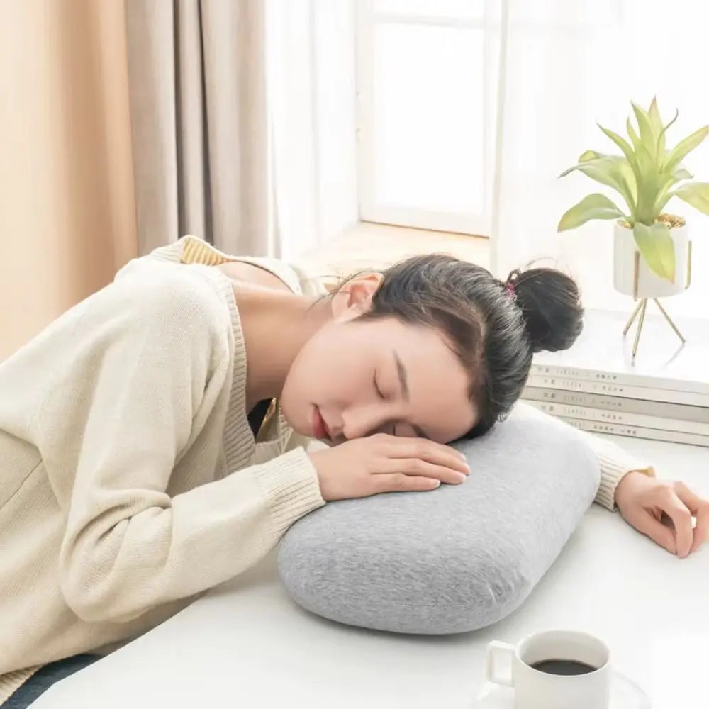 A person is resting their head on a gray memory foam travel neck pillow indoors near a window.