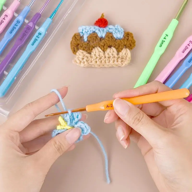 A hand is using an orange crochet hook to work on a blue and brown crocheted cake decoration.