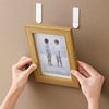 Hands are hanging a wooden picture frame on a wall using removable adhesive picture hanging strips.