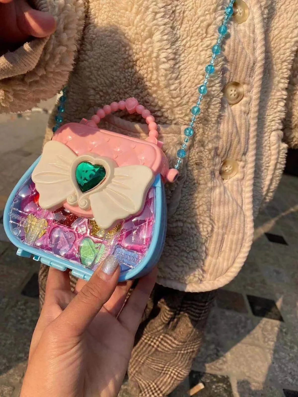 A hand holds a pink and white makeup toy set with a heart-shaped butterfly detail and a blue beaded strap.