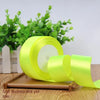 A roll of bright fluorescent yellow satin ribbon lying on a wooden surface with green foliage in the background.