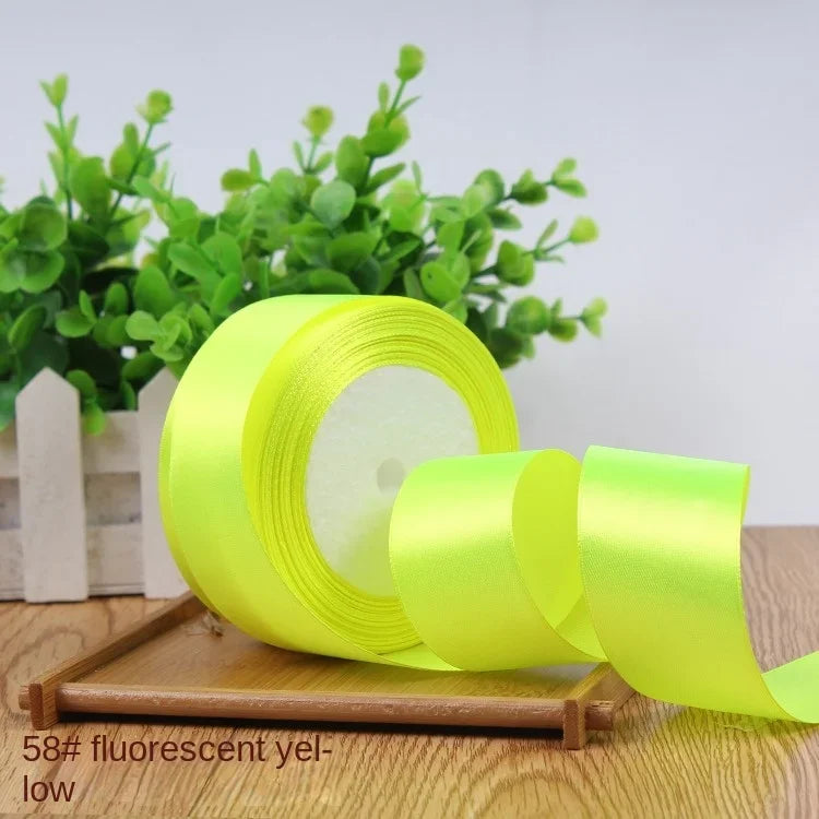 A roll of bright fluorescent yellow satin ribbon lying on a wooden surface with green foliage in the background.