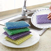 A collection of four colorful double-sided kitchen scouring sponges stacked on a white plate next to a kitchen sink.
