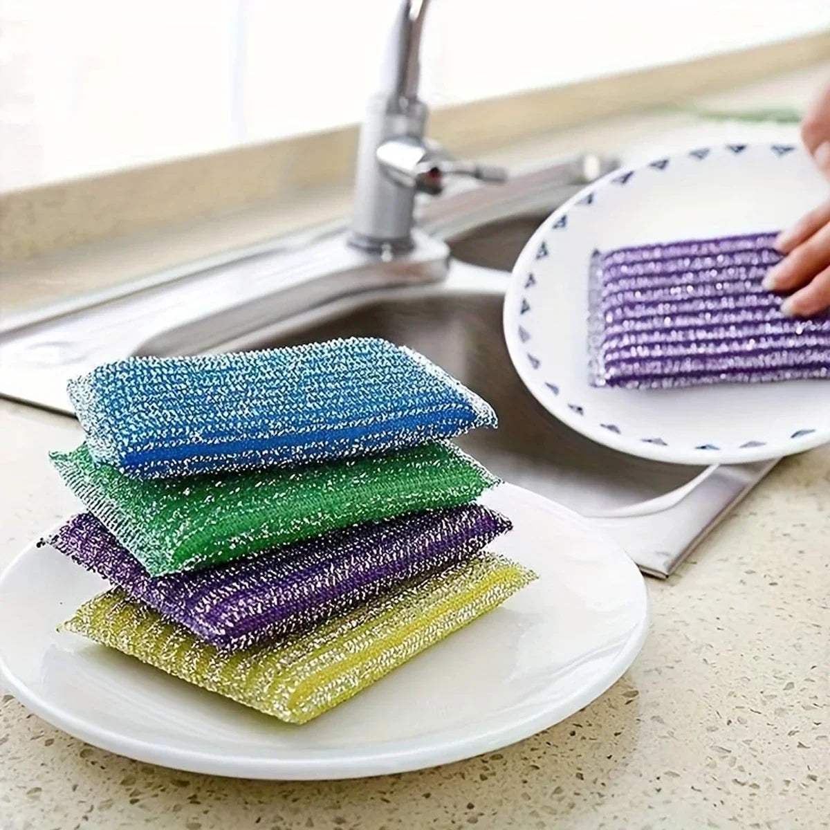 A collection of four colorful double-sided kitchen scouring sponges stacked on a white plate next to a kitchen sink.