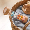 A glass bottle filled with colorful pom-poms and wooden rattle components inside a woven basket.