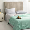 A light mint green cooling summer blanket is displayed on a bed with a neutral upholstered headboard.