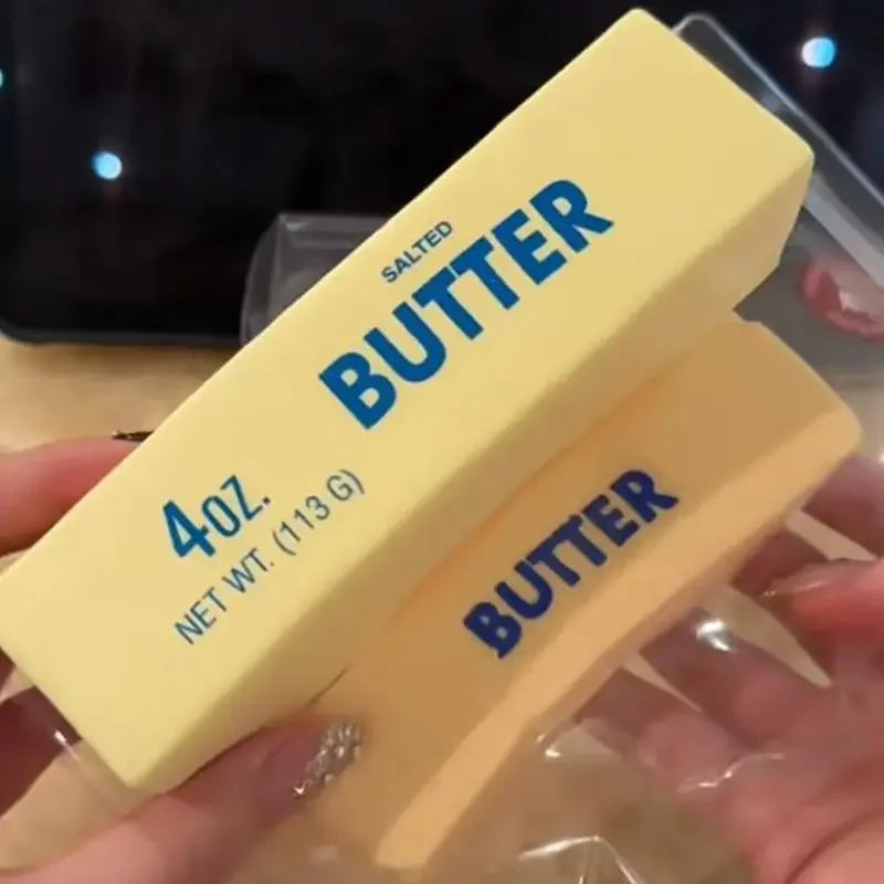 A hand holds a yellow squishy butter stick fidget toy with blue lettering, showing its size and texture.