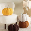 A yellow pumpkin plush pillow sits on a white chair next to a dark brown pumpkin cushion in a bright room.