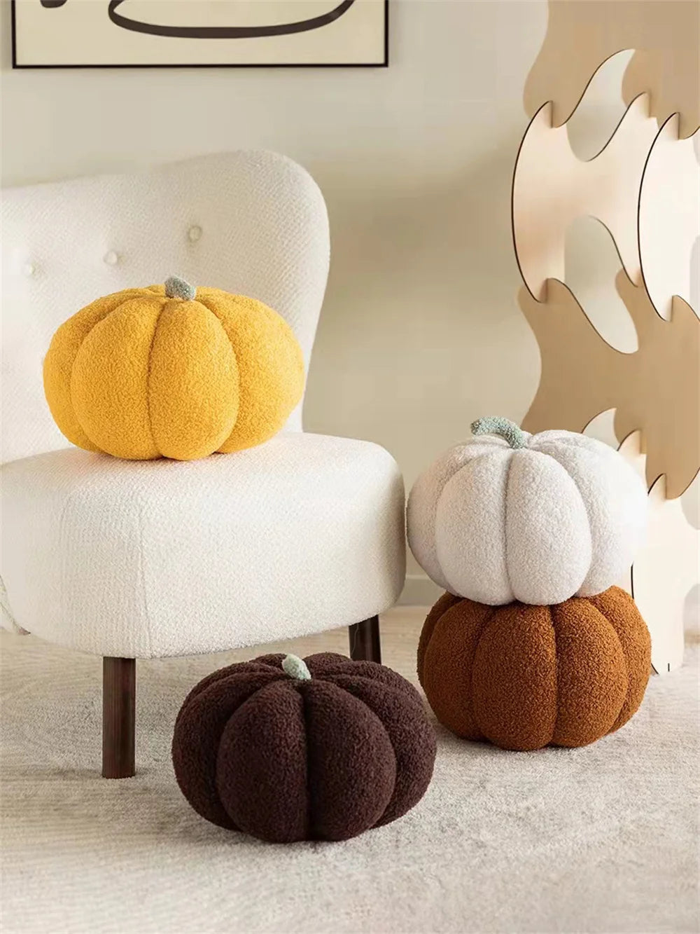 A yellow pumpkin plush pillow sits on a white chair next to a dark brown pumpkin cushion in a bright room.