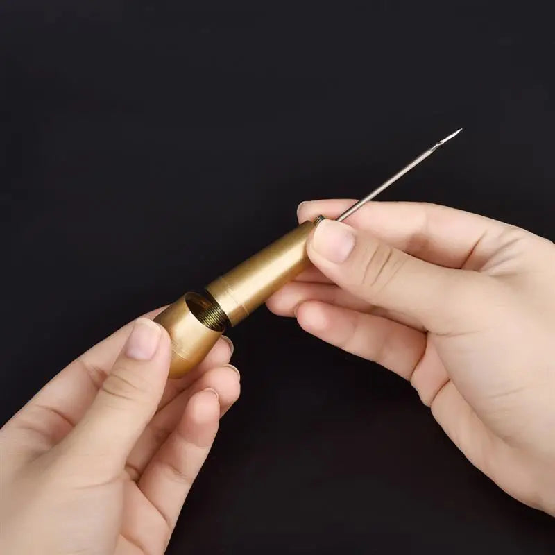 A hand holds a gold-colored leather sewing awl set with a replaceable needle.