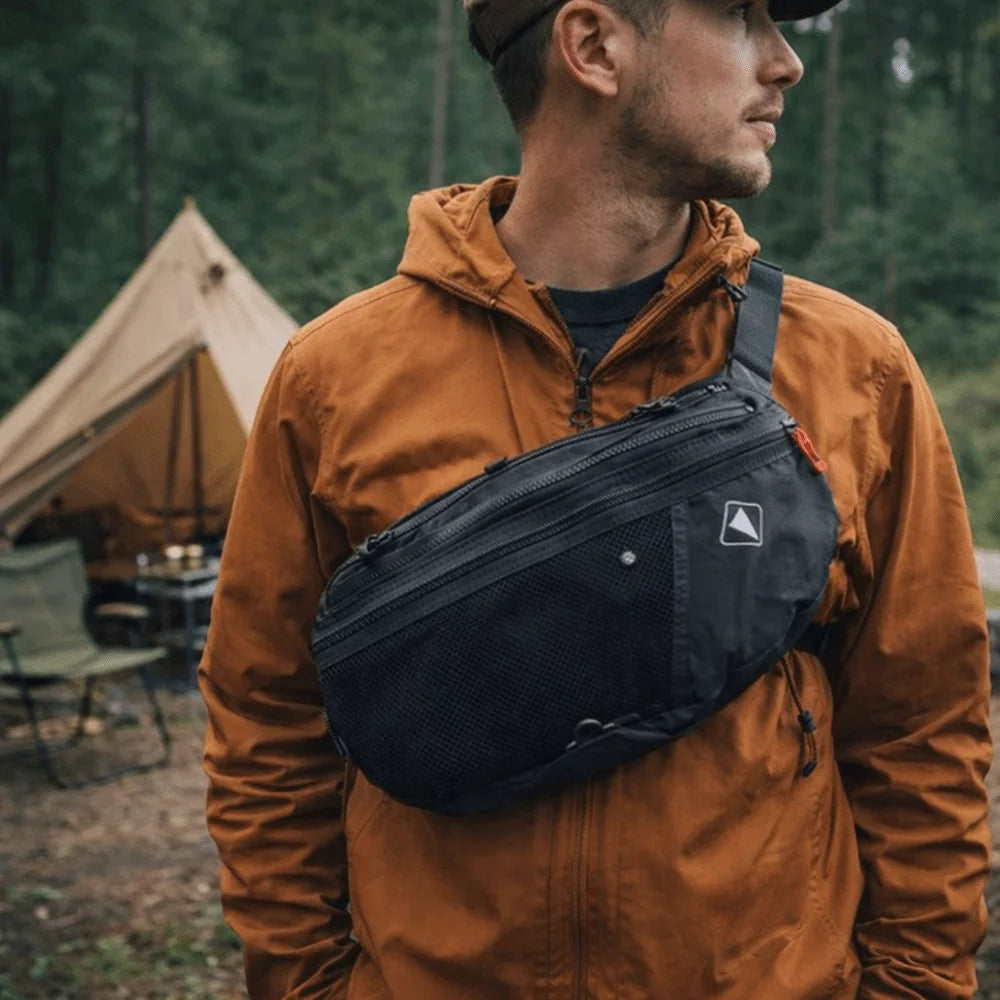 A man wearing an orange jacket holds a black nylon crossbody bag with an adjustable sling strap.