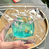 A hand holds a clear plastic bag containing a turquoise ocean-themed stress relief squeeze toy with seashell decorations.