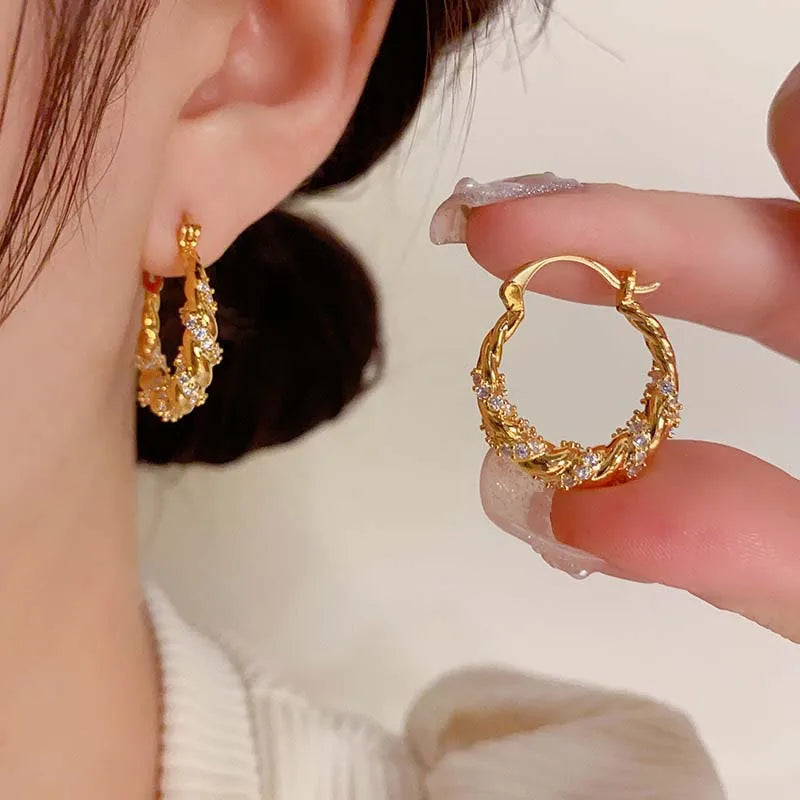 Gold twisted zircon hoop earrings displayed on a hand against a blurred background.