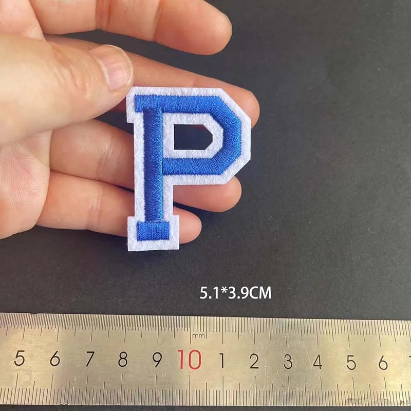 A hand holds a blue embroidered letter P patch, measuring 5.1 by 3.9 cm against a ruler.