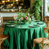 A round table set with a green satin tablecloth, place settings, and floral decorations in a bright indoor setting.
